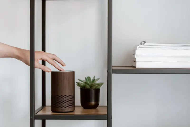 A hand interacts with a smart speaker on a minimalist shelf, featuring a plant and stacked books