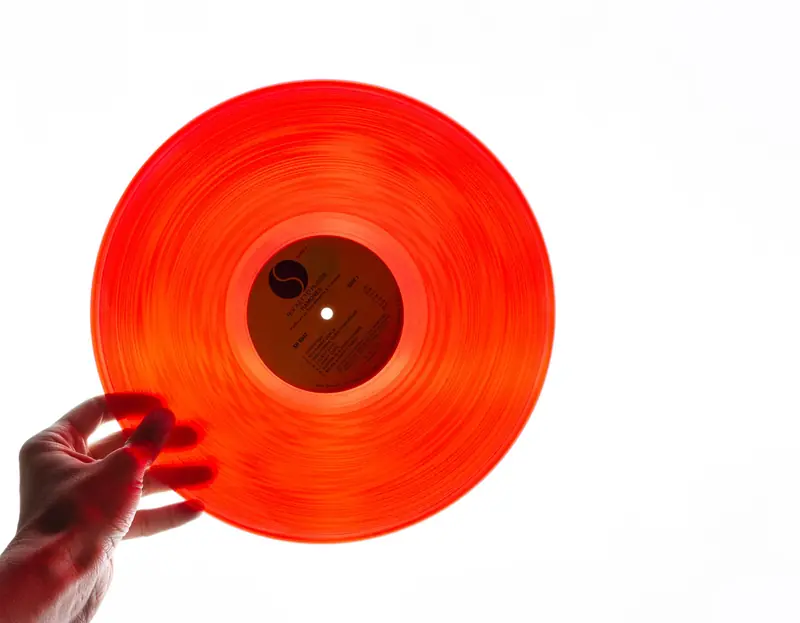 A bright orange vinyl record being held by a hand against a plain white background, showcasing its translucency
